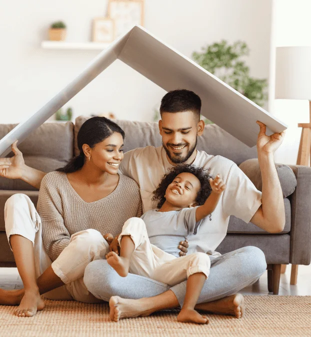 A family of three, with the father holding a cardboard roof over his family’s head.