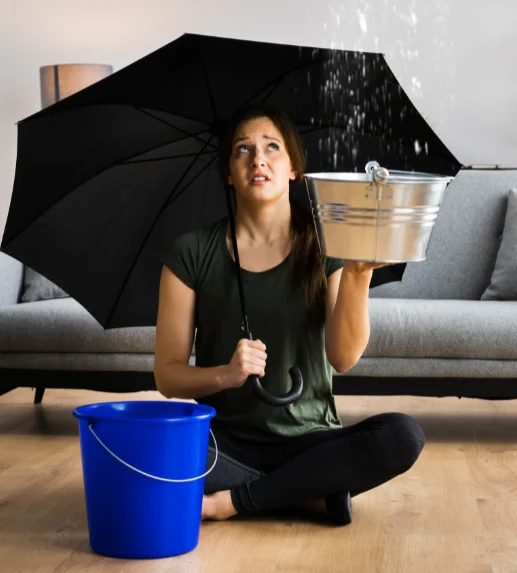 woman sitting on floor holding black umbrella and an iron bucket next to a blue bucket with a large sofa behind her